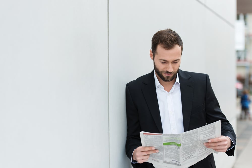 Businessman reading a newspaper on his lunch break as he leans against a white wall with copyspace